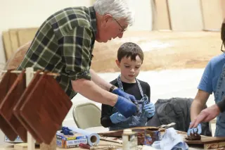 Volunteer helps a child apply finish to wooden boat parts at a workshop table