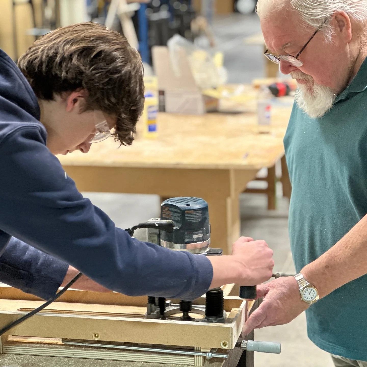 Adult instructor guides a teenager using a handheld router on a wooden project.
